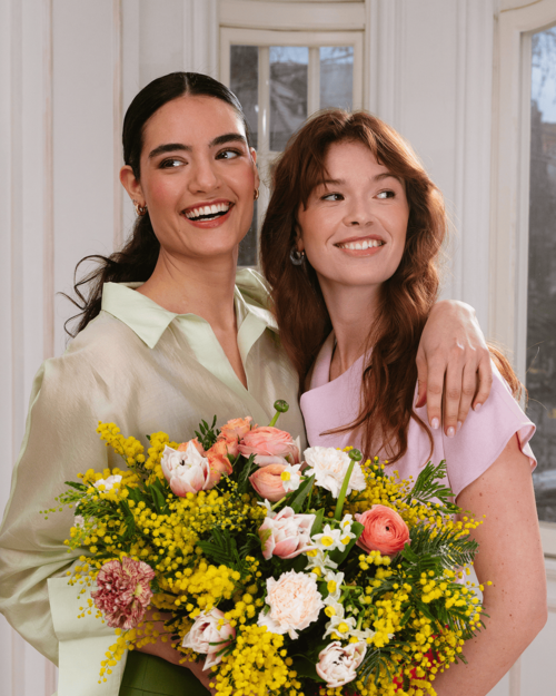 Due donne sorridenti abbracciate con bouquet di mimosa e fiori primaverili tra le mani, in un ambiente luminoso