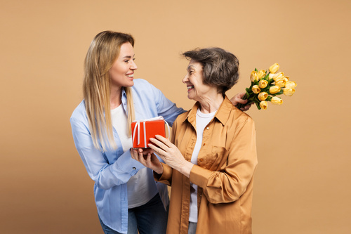 Figlia porge un regalo rosso alla madre sorridente che tiene un bouquet di fiori gialli.