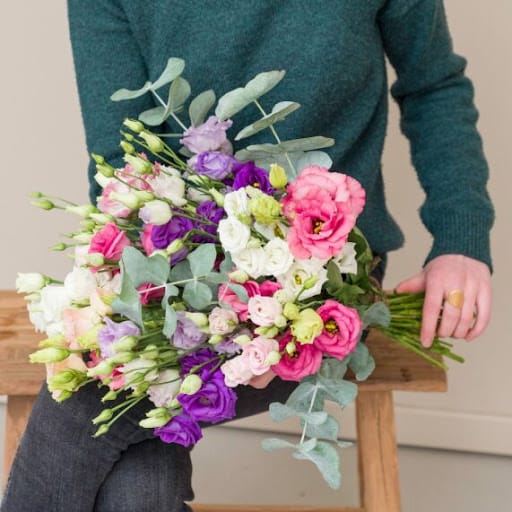 bouquet di lisianthus bianchi, rosa e viola con decorazioni verdi tenuto in mano da una persona seduta su una panca